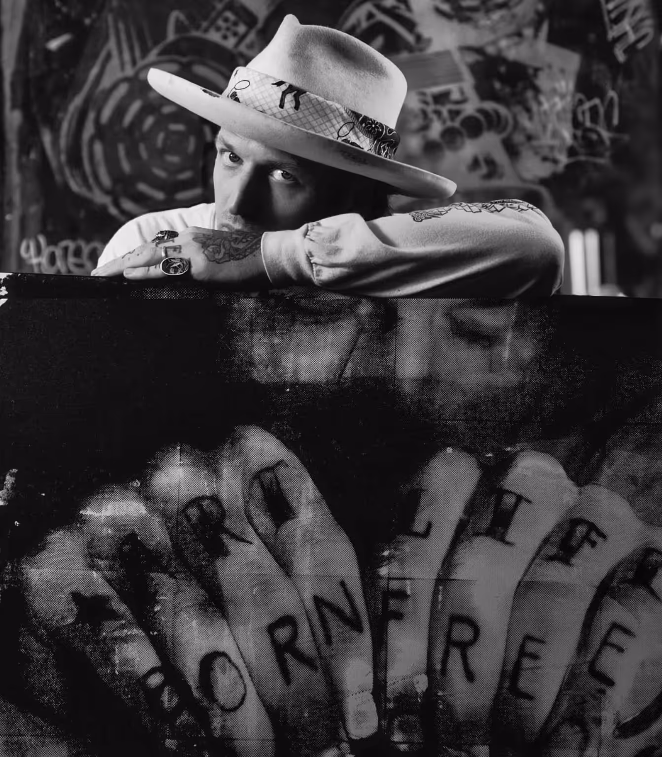 Black-and-white portrait of tattooed man in a hat resting arms on a canvas with tattooed hands image.