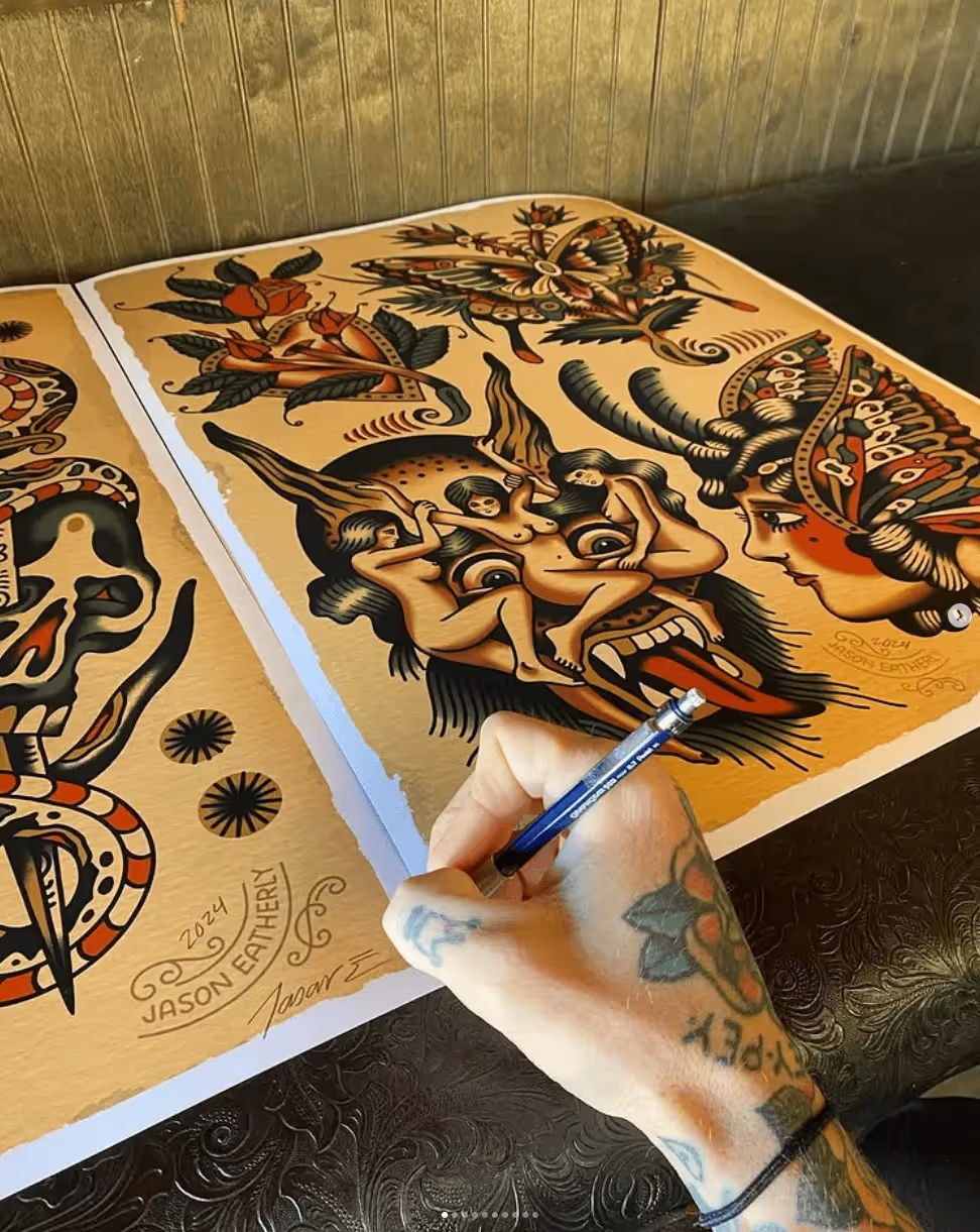 Artist's tattooed hand signing a print of vintage-style tattoo art featuring figures and a butterfly.