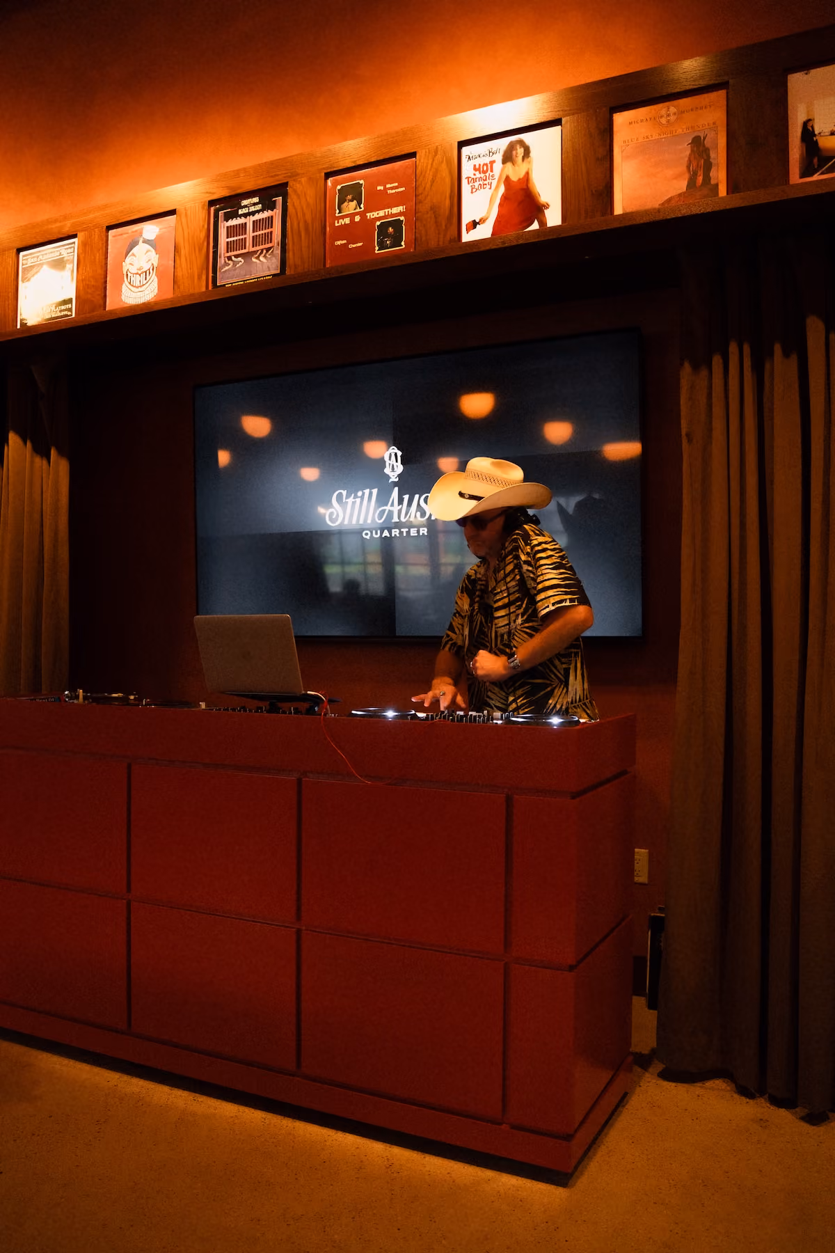 A DJ in a cowboy hat and patterned shirt mixes at a red DJ booth in the Vinyl Lounge, with a Still Austin Quarter screen and a shelf of vintage album covers behind him.