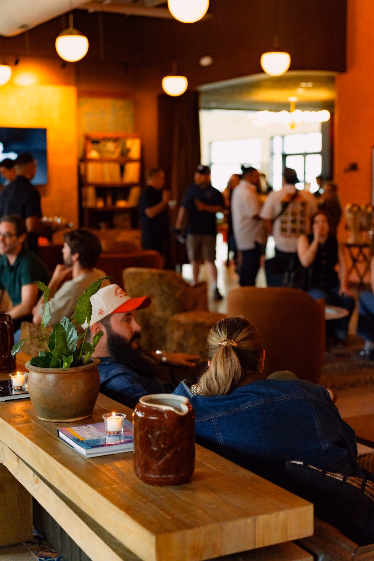Guests relax and talk in the Still Austin Quarter lounge, with candles and a potted plant on a wooden table in front and globe pendant lights overhead.