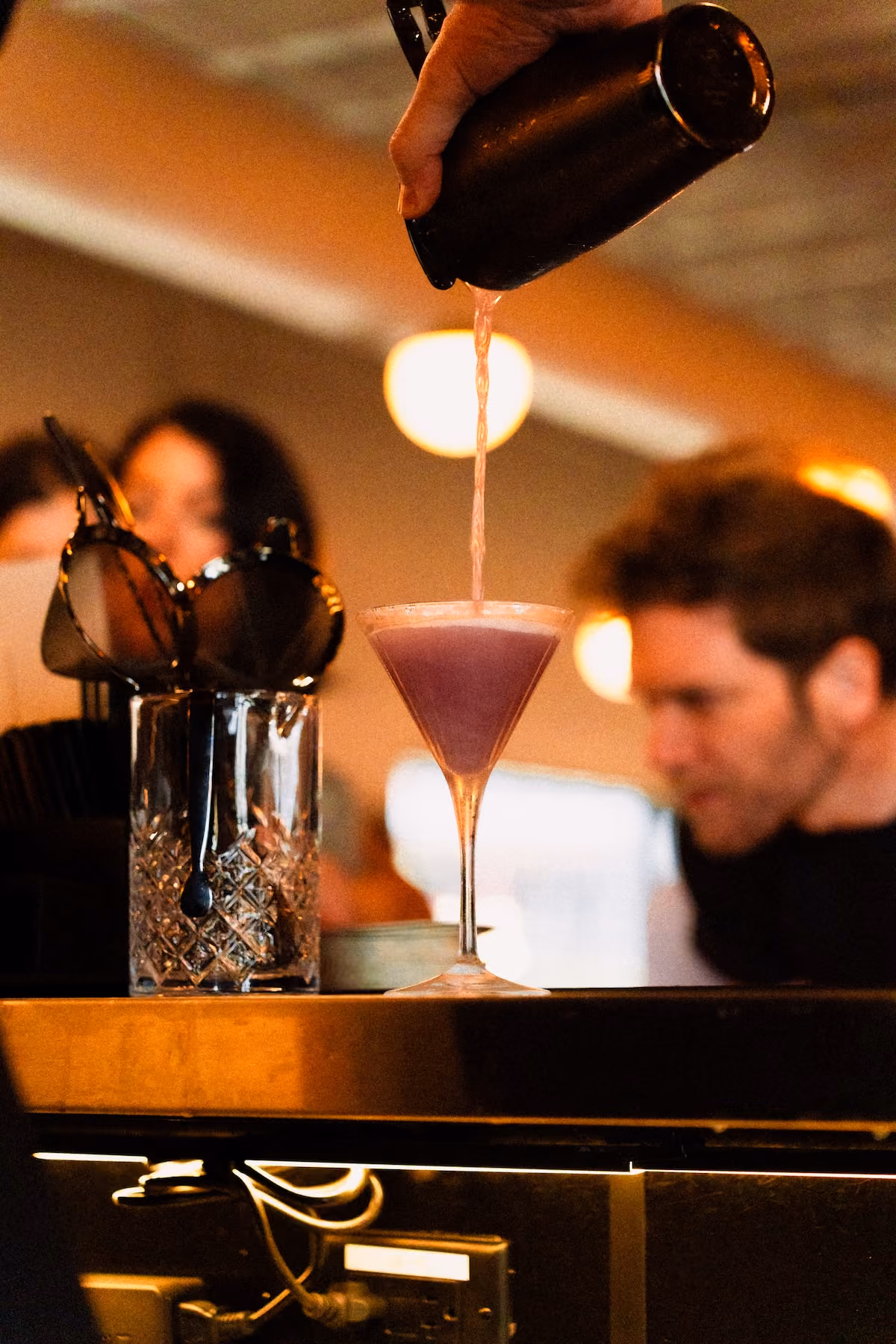 A bartender pours a pink cocktail from a shaker into a martini glass at the Still Austin Quarter bar, with warm pendant lights and guests softly blurred in the background.
