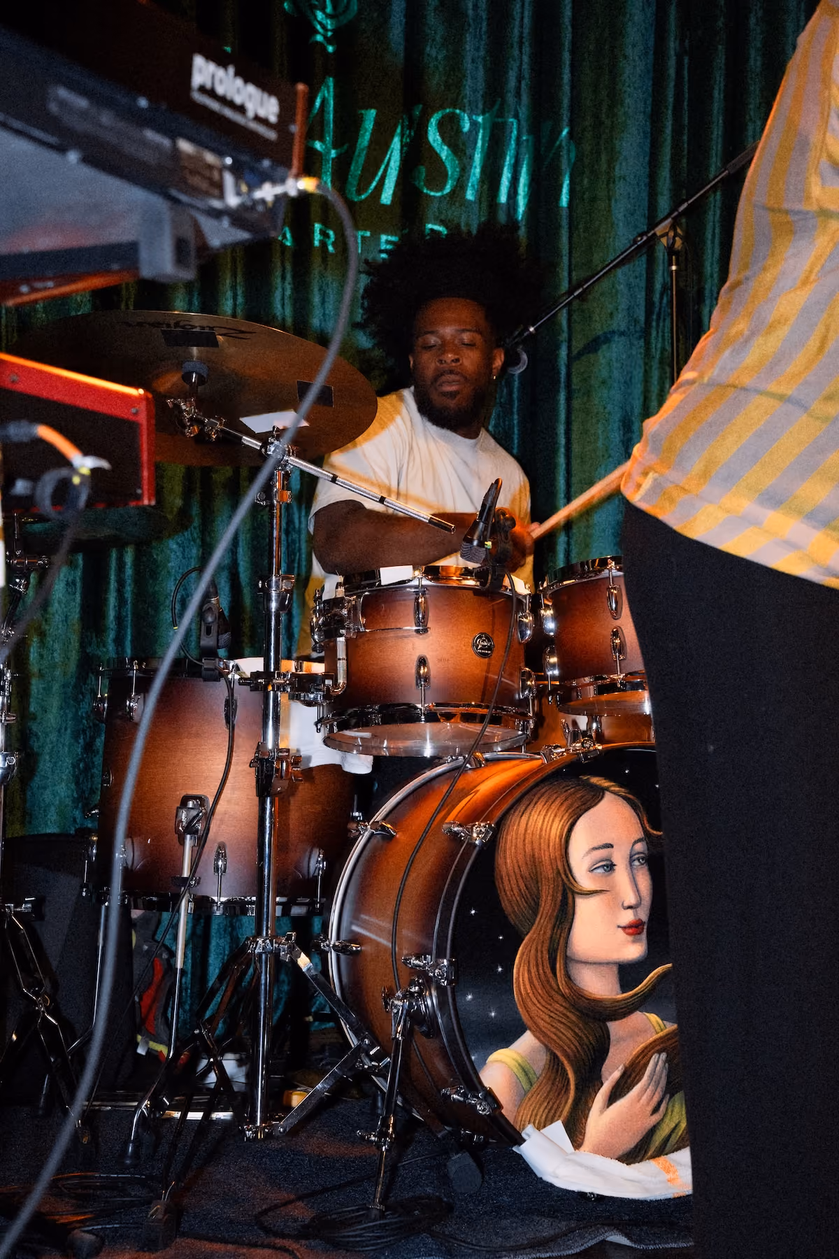 A drummer plays a live set on The Musician’s Stage in the Music Room, in front of a green velvet curtain with the Still Austin Quarter name, his bass drum painted with a portrait of a woman.