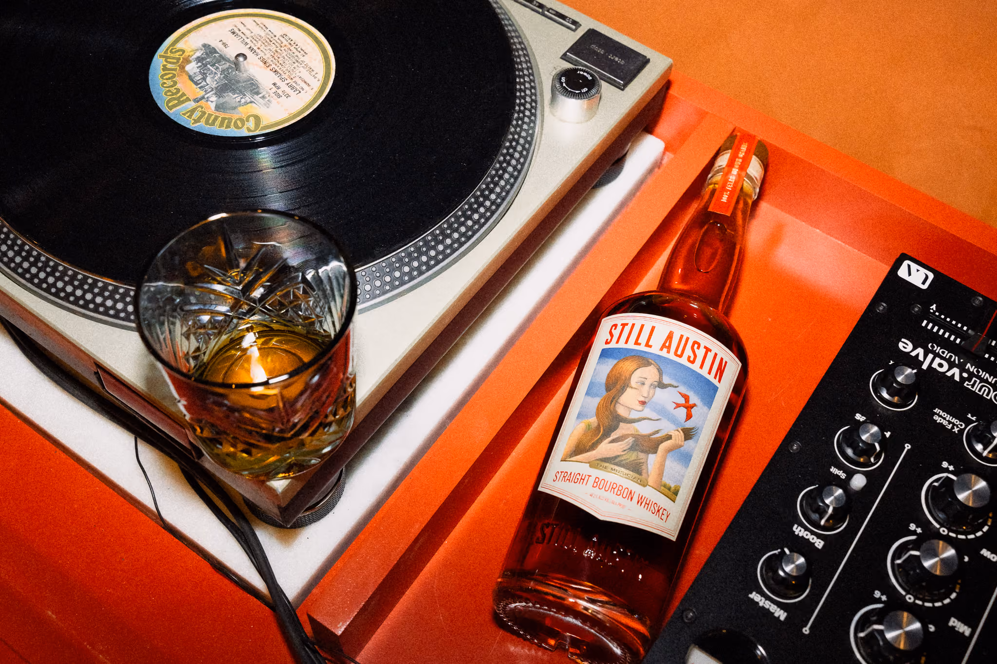 A glass of whiskey and a bottle of Still Austin The Musician bourbon beside a turntable