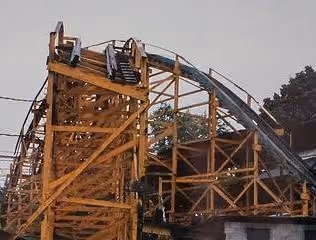 Wooden roller coaster structure with curved track and stairs against a pale sky.