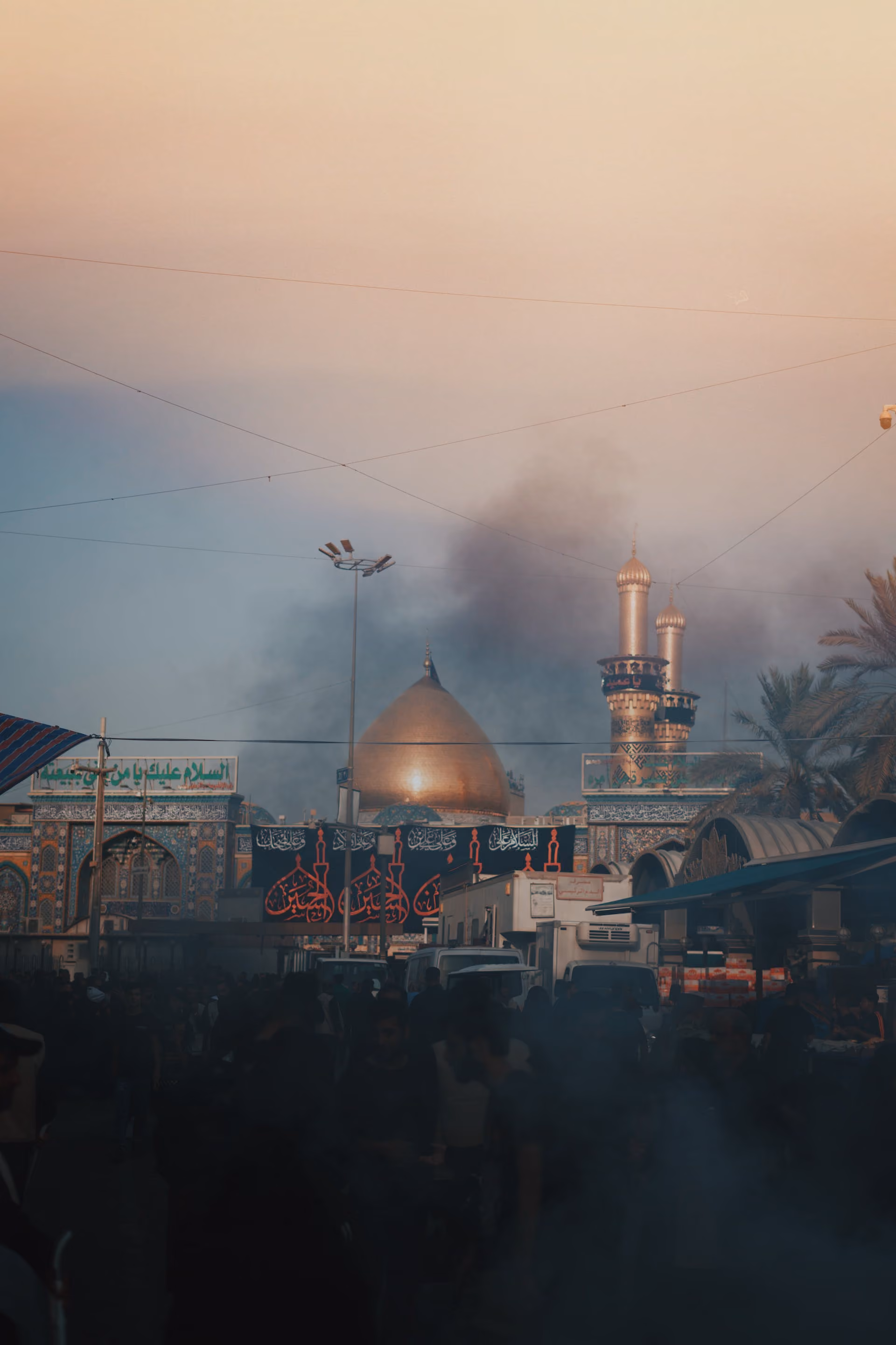 Crowd and vehicles below golden-domed mosque with minarets and smoke in the sky at sunset.