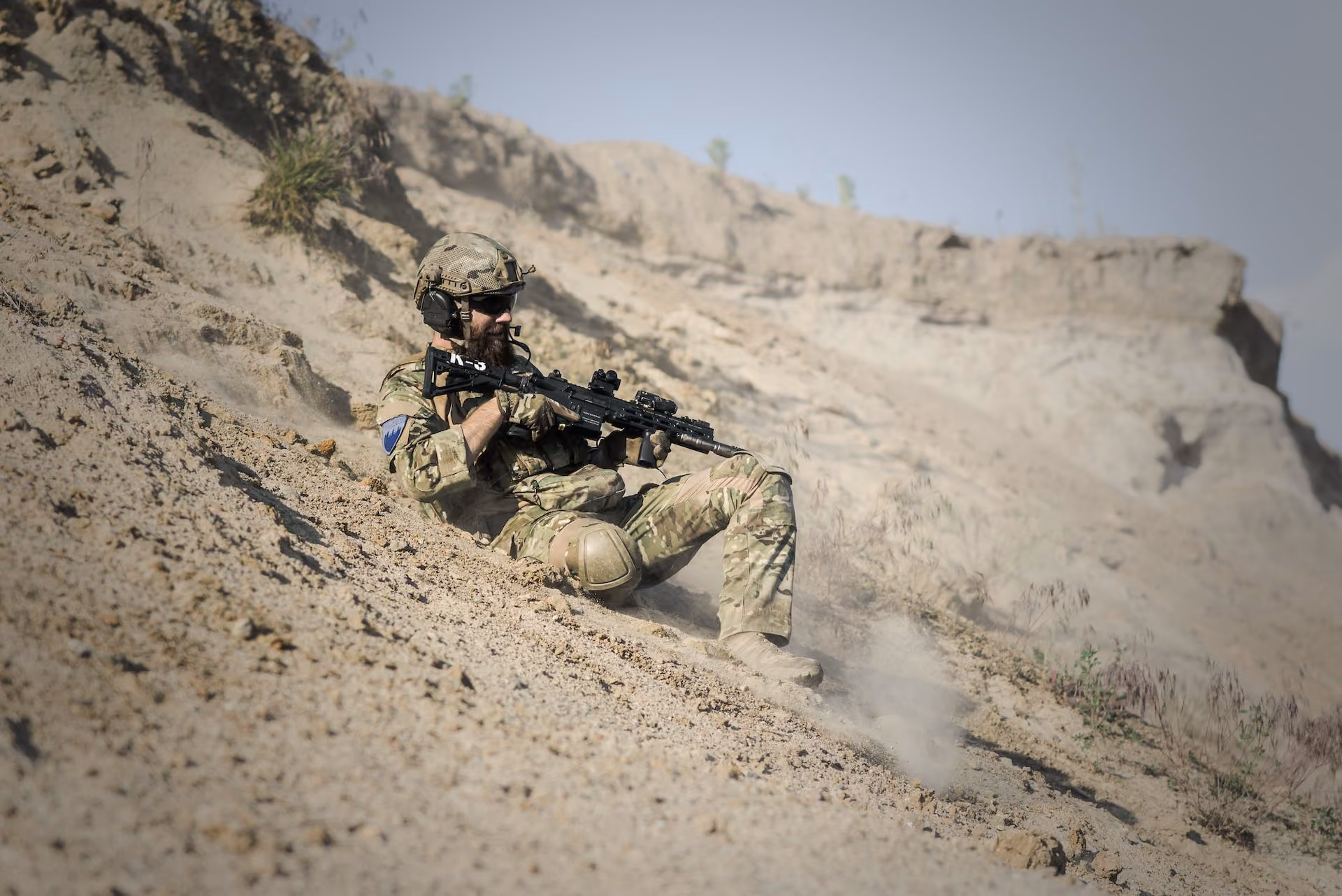 Soldier in camouflage gear sliding or falling down a sandy slope holding a rifle.