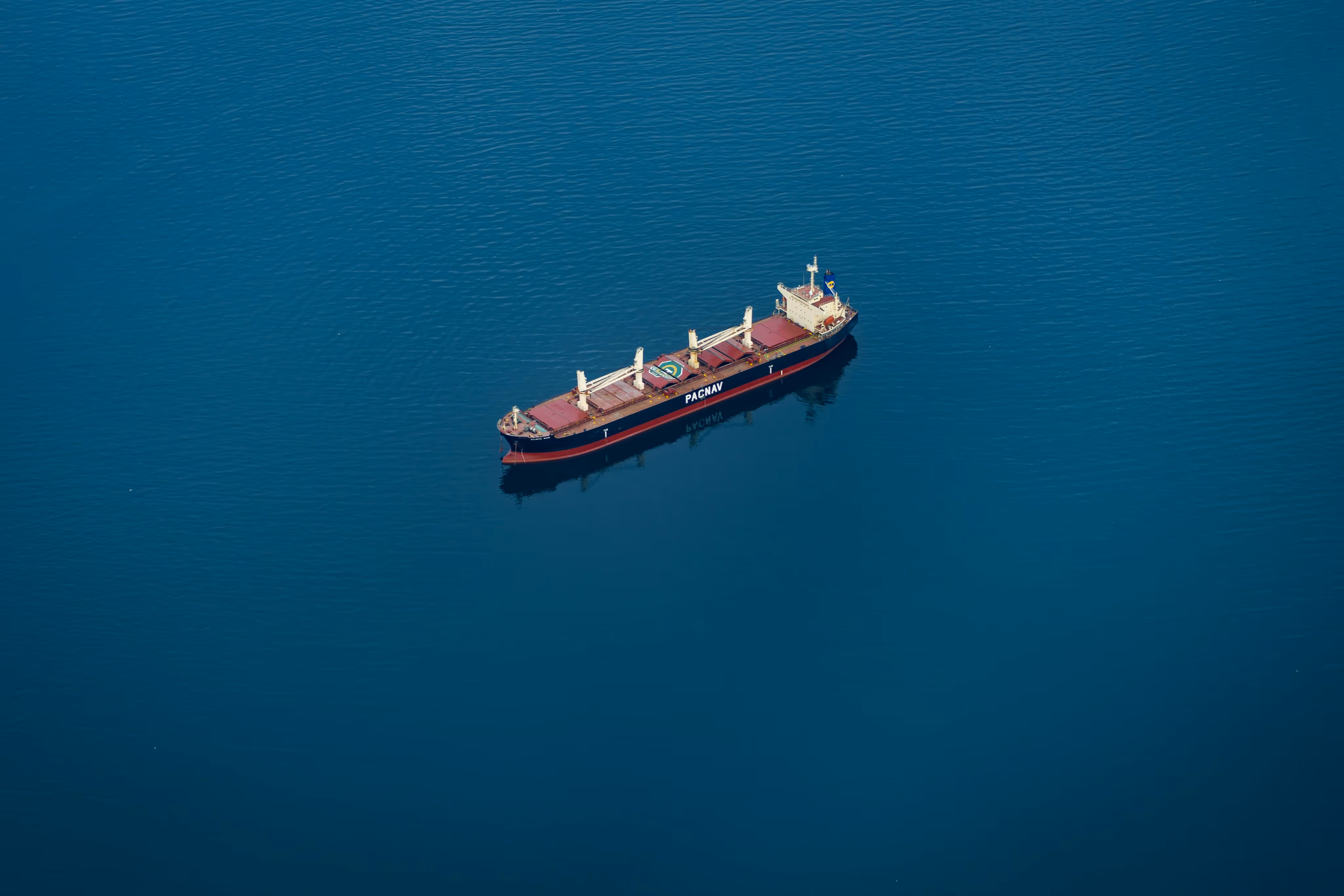 Large cargo ship named PACNAV floating on calm deep blue water viewed from above.