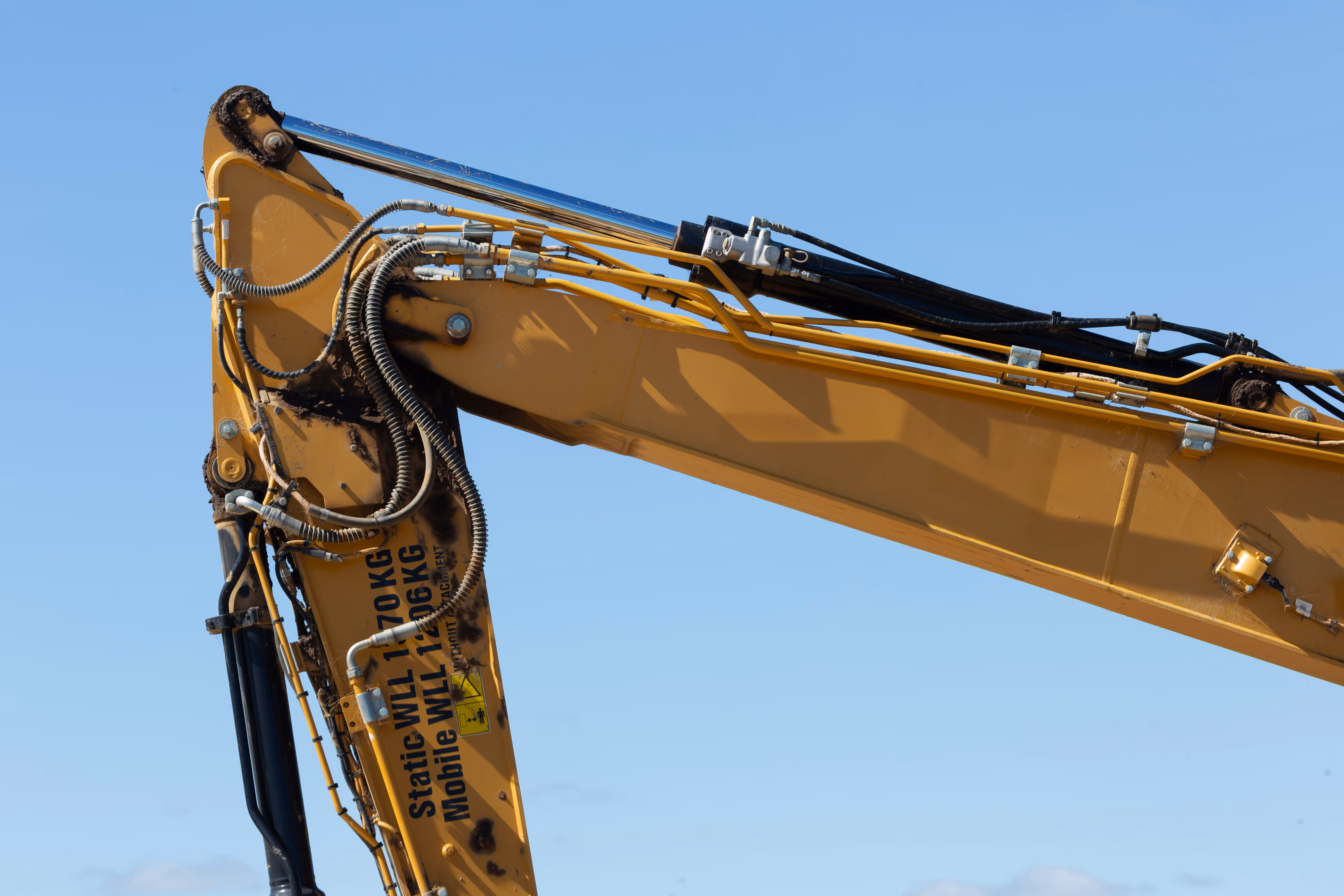 Close-up of a yellow excavator arm with hydraulic pipes against a blue sky.