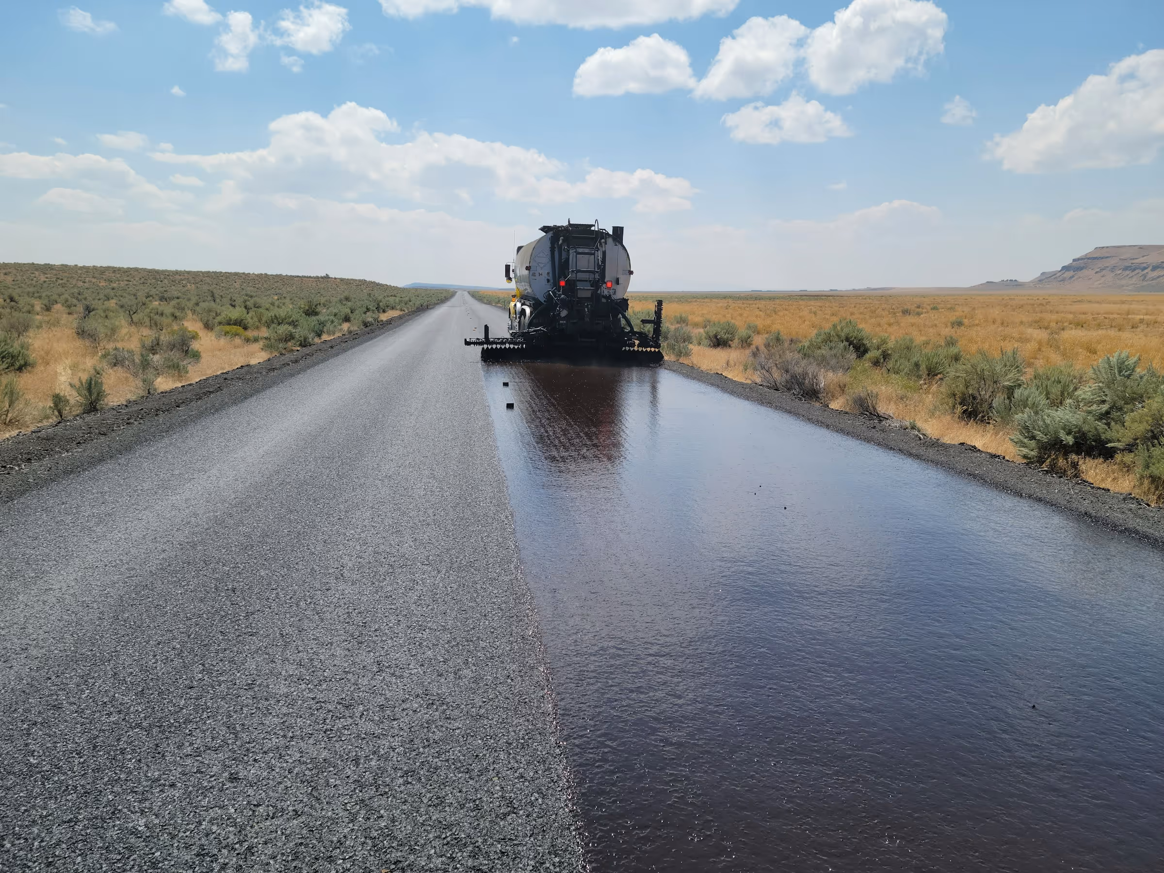 Machine applying liquid asphalt on a rural road under a partly cloudy sky.