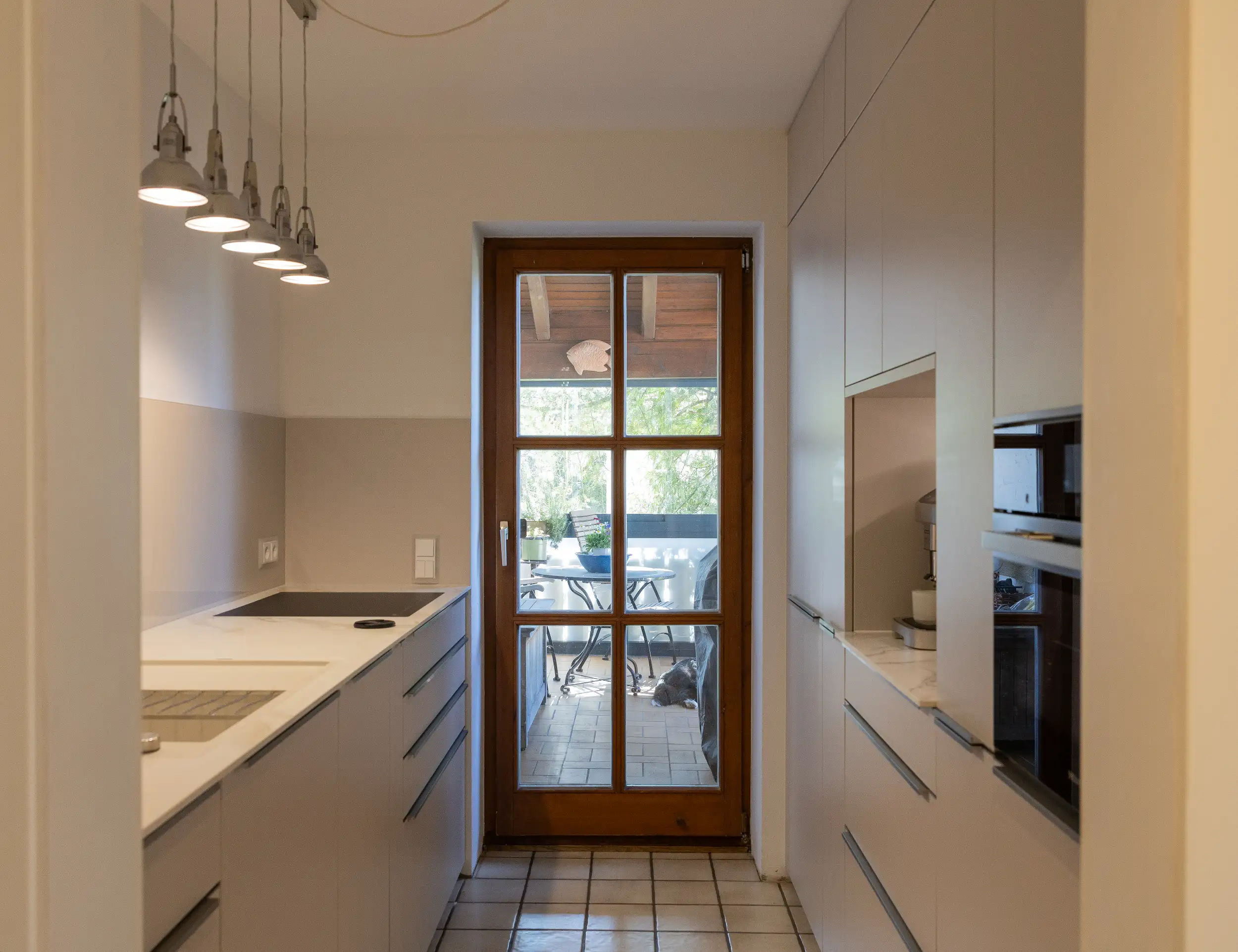 Auf dem Bild sieht man eine Küche mit Sahara Melaminharz Fronten, Griffleisten in Metall Optik, marmorierter Keramik und Nischenpanel in Glas. Deckenhohe Küchenschränke, Premium Auszüge und hochwertige Geräte sorgen für genug Stauraum und echten Komfort. Geplant vom Thoki Küchenstudio in Wörthsee für anspruchsvolle Kunden aus München, Starnberg und Umgebung. 