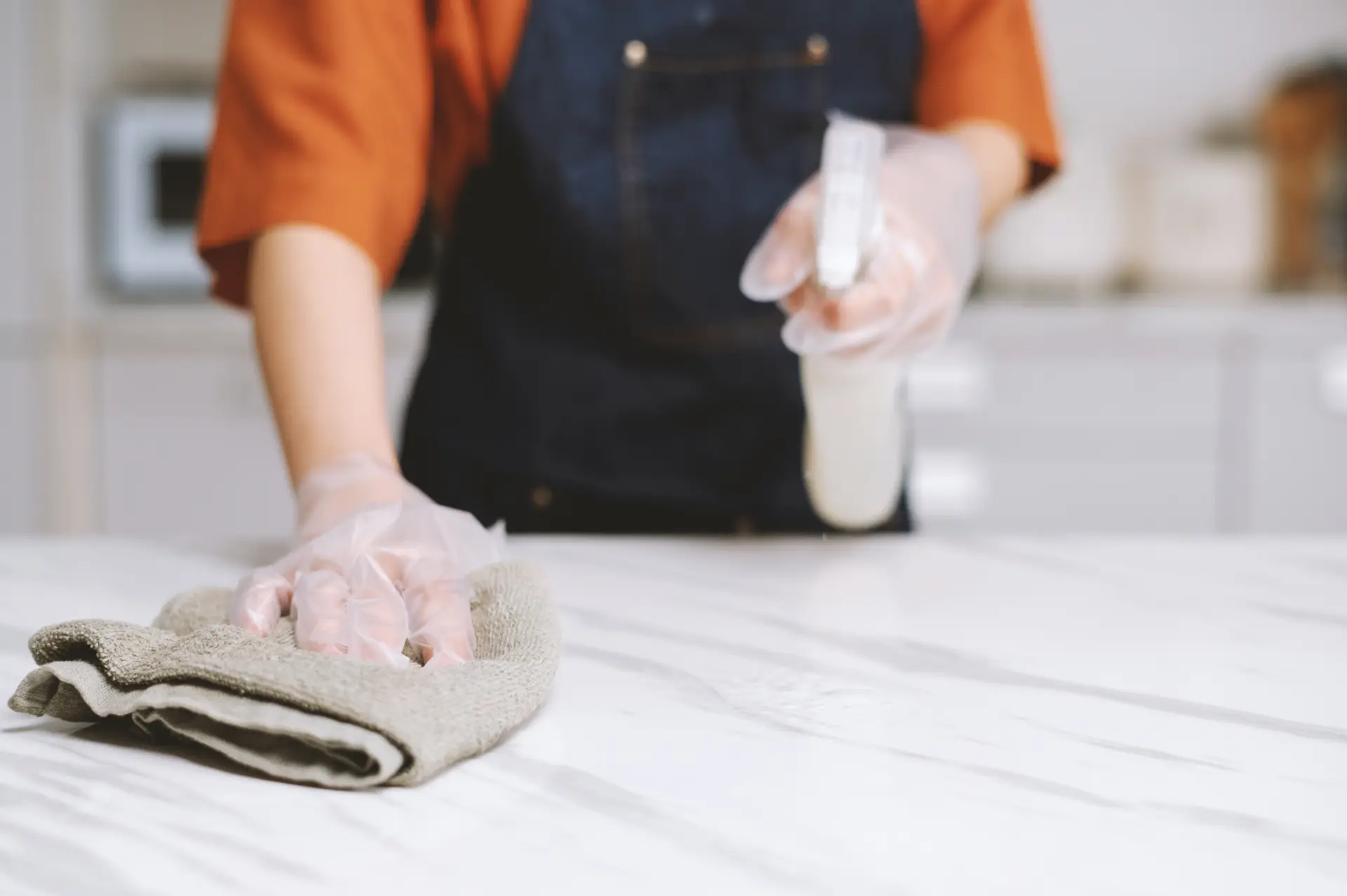 Person reinigt eine weiße Marmor-Arbeitsplatte mit Mikrofasertuch und Sprühreiniger, trägt dabei durchsichtige Handschuhe – Küchenarbeitsplatte richtig reinigen ohne Rückstände