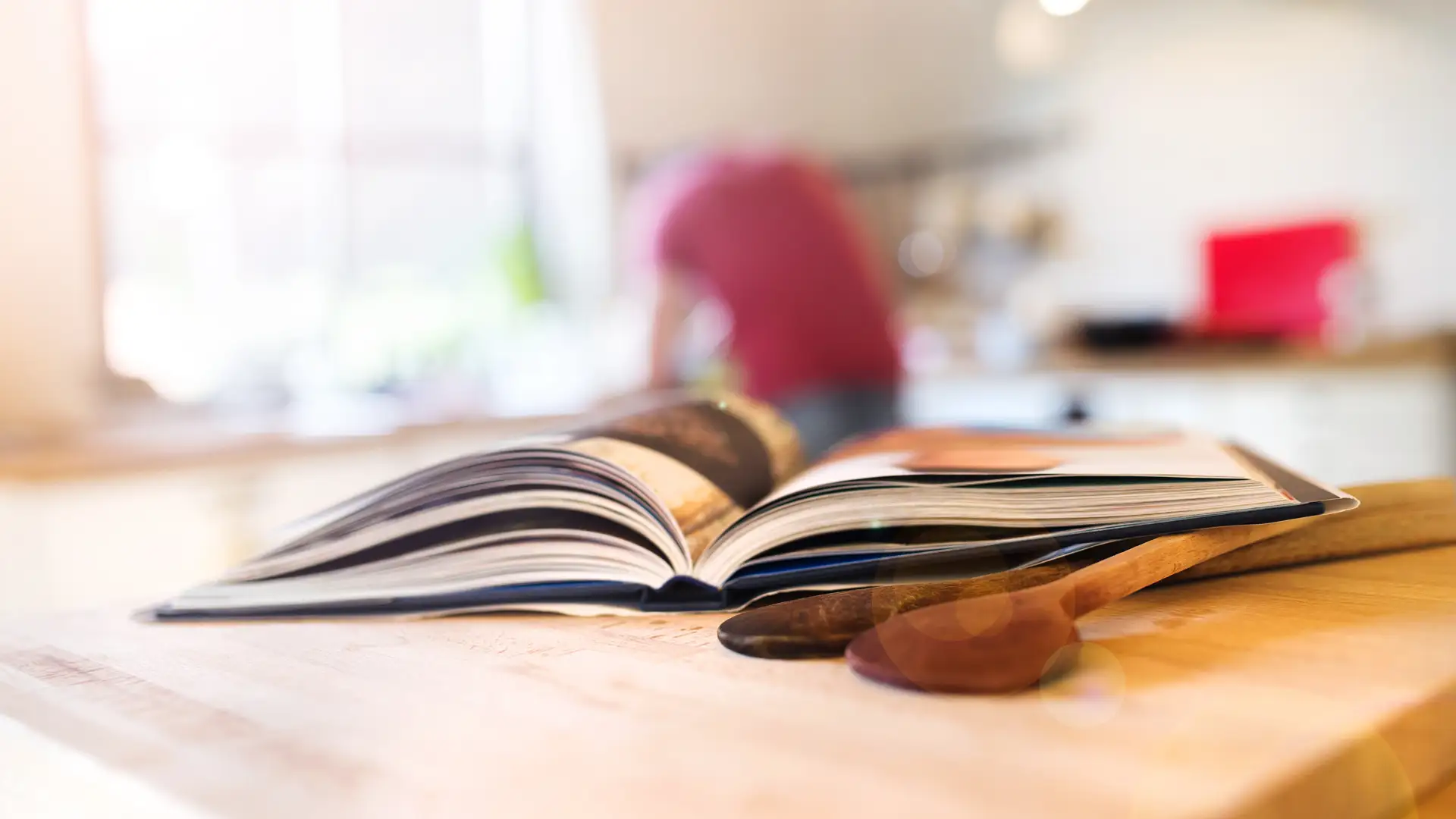 Aufgeschlagenes Kochbuch und Holzlöffel auf Arbeitsplatte mit unscharfem Hintergrund