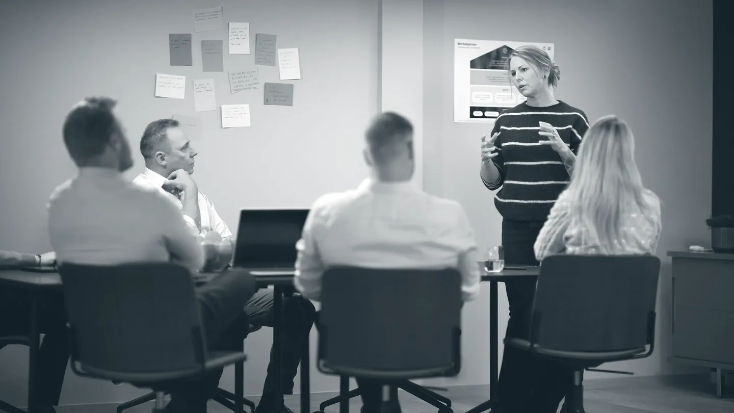 Woman standing and speaking to a group of four seated people in a meeting room with sticky notes on the wall.