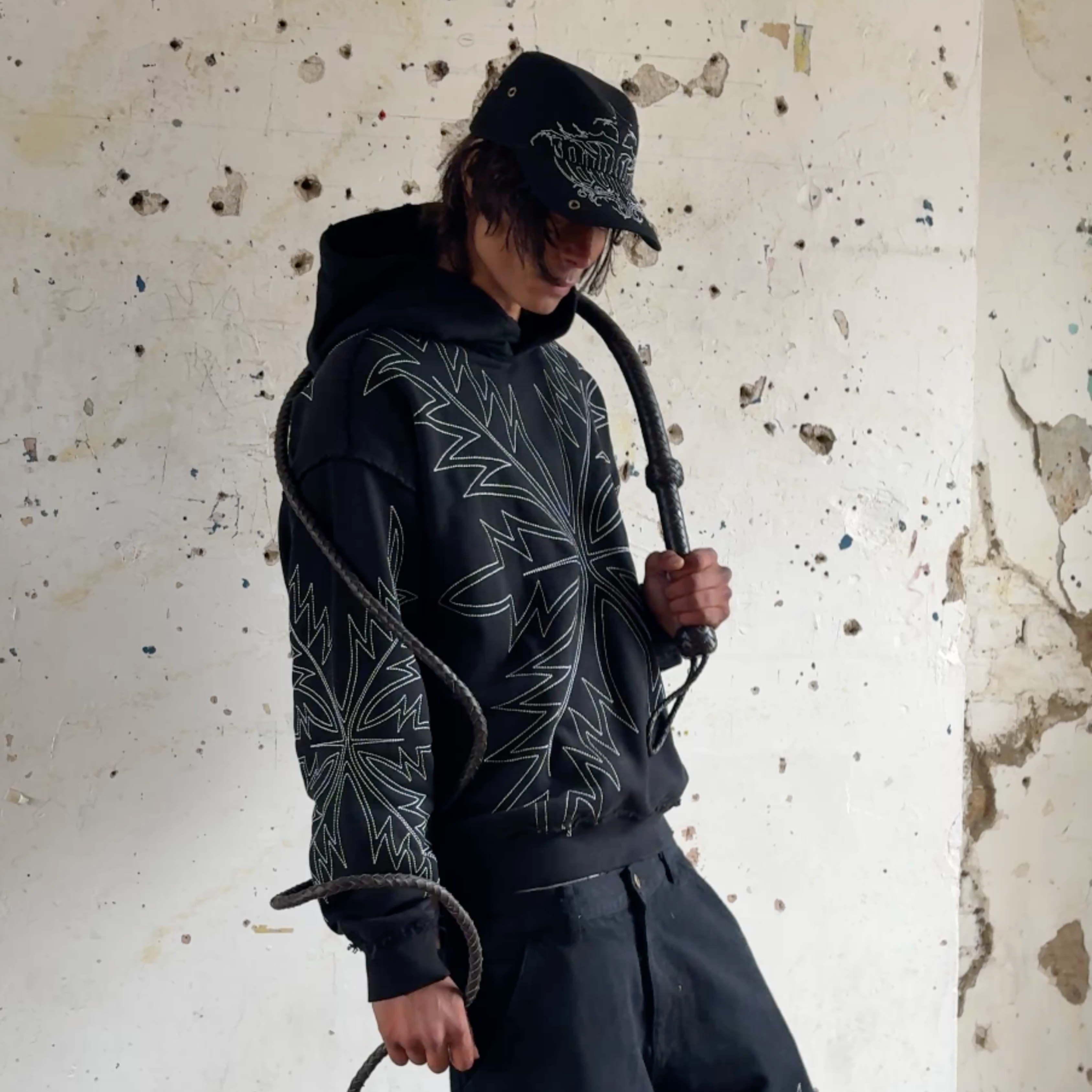 male model wearing western stitch hoodie and streetwear cowboy hat whilst holding whip 
