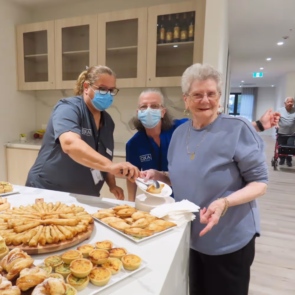 Elderly woman being served pastries by staff in masks at our aged care home.