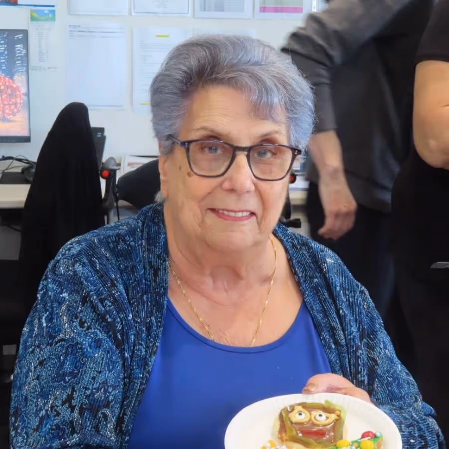 Ida Cinerari, an elderly woman with short grey hair and glasses, smiling while holding a decorated cookie on a plate during a creative activity at Oxa Care.