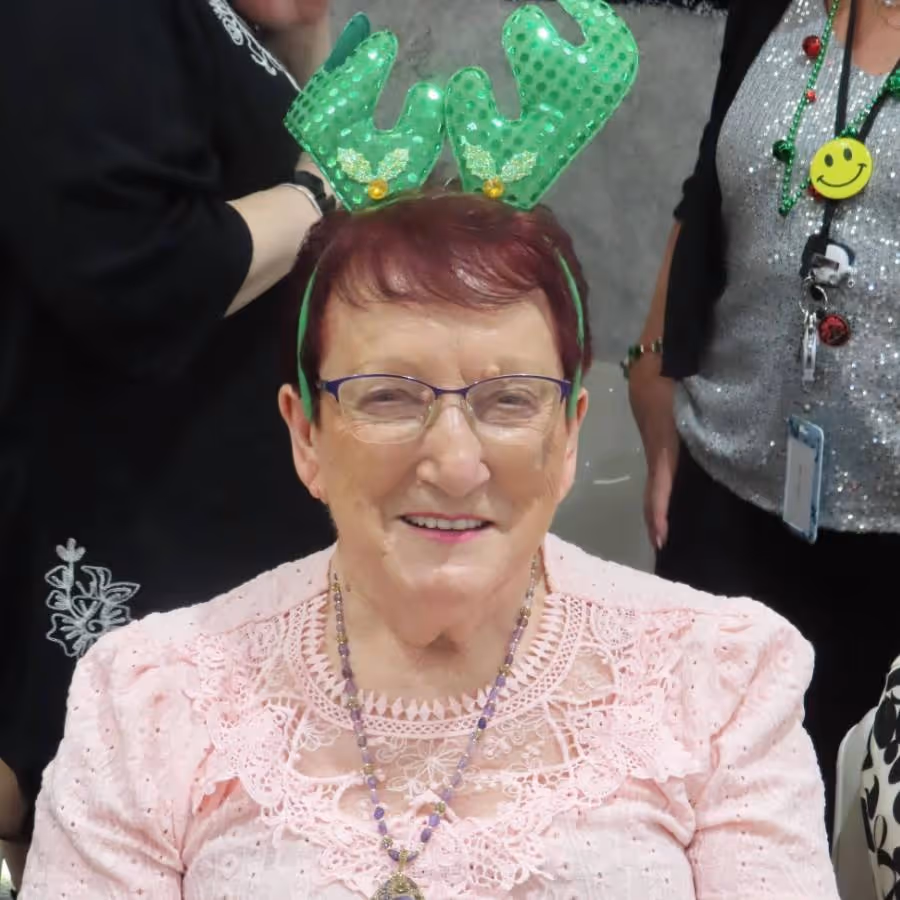 Verna Stubberfield, an elderly woman with short red hair and glasses, smiling while wearing green reindeer antlers during a festive celebration at Oxa Care.