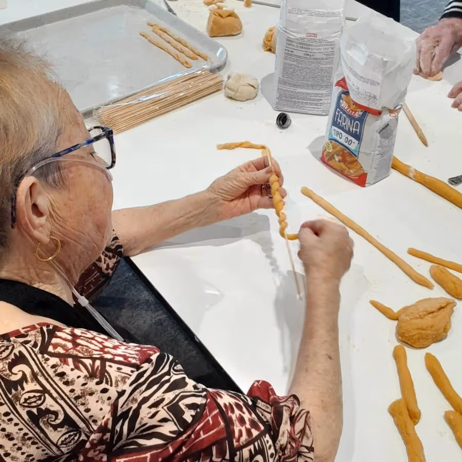 Melina Caporetto, an elderly woman wearing glasses and an apron, carefully shapes homemade pasta dough by hand during a cooking activity at Oxa Care.