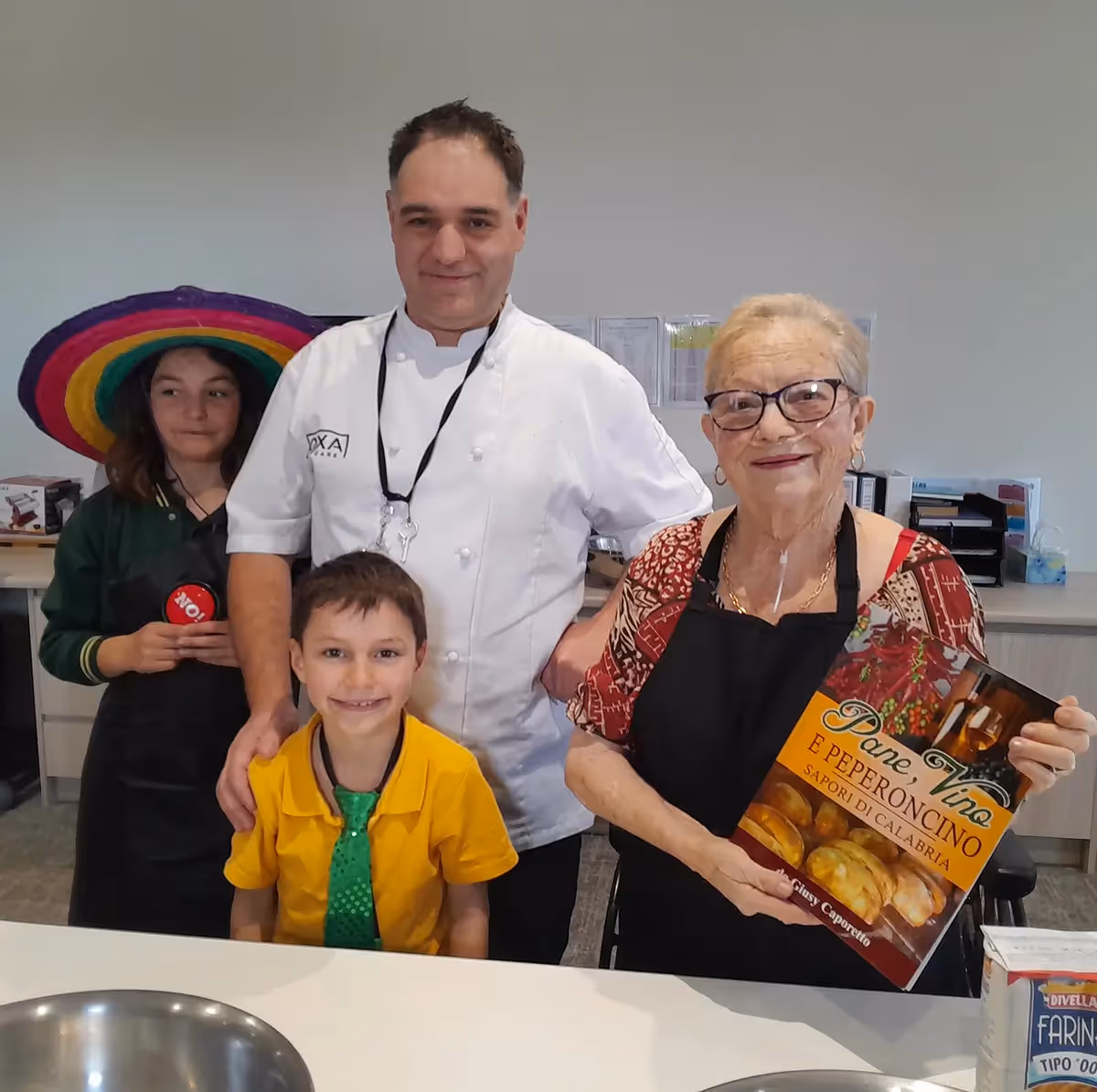 Smiling Italian woman holding a cookbook alongside a chef and children during a cooking activity.