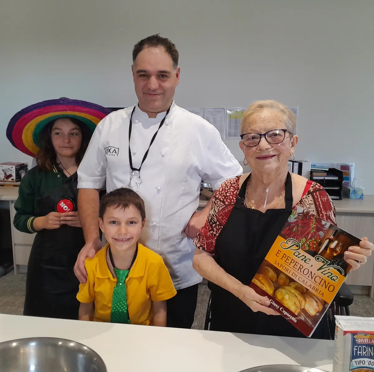 Smiling Italian woman holding a cookbook alongside a chef and children during a cooking activity.