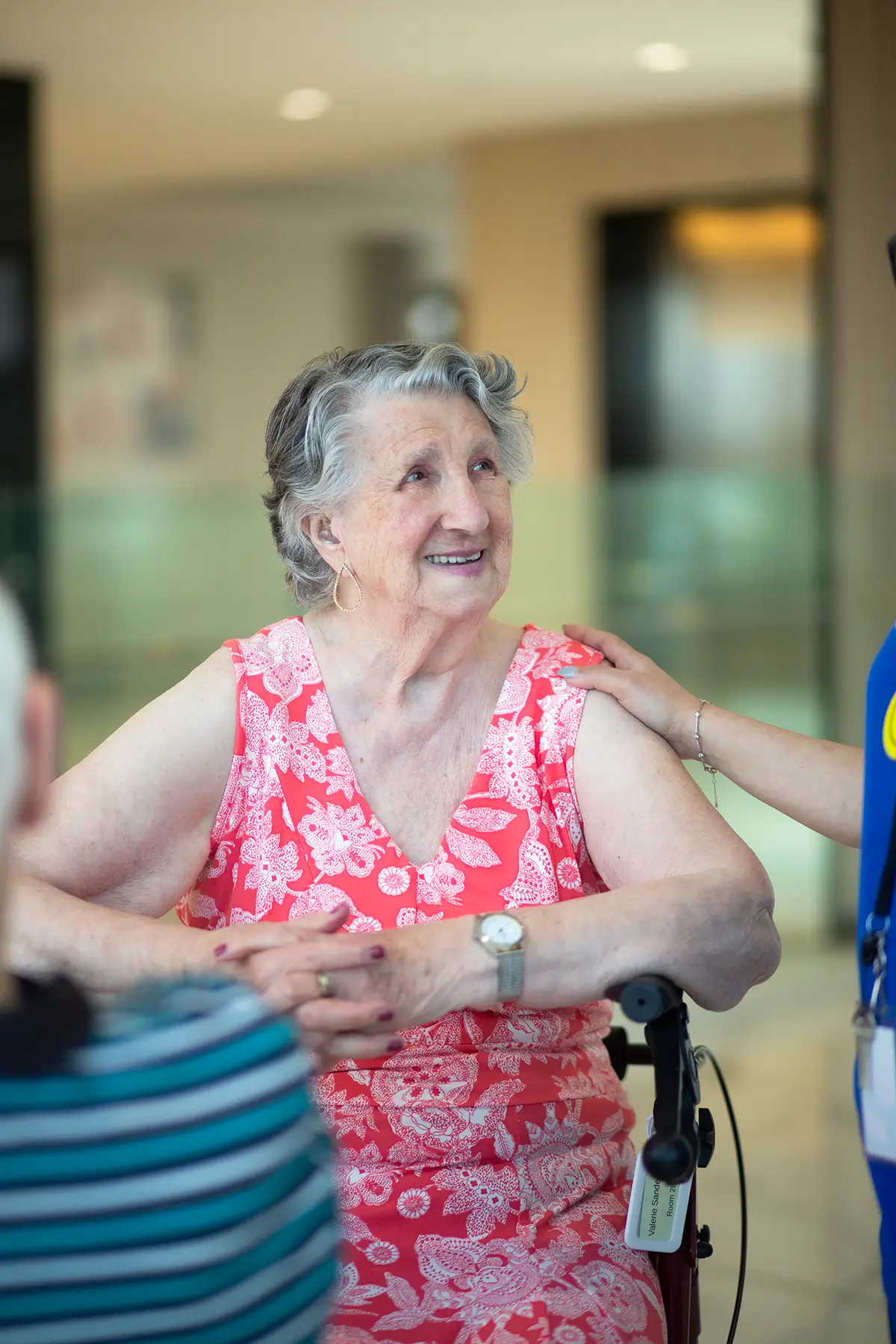 Smiling elderly Italian woman in a pink floral dress receiving support from a caregiver.