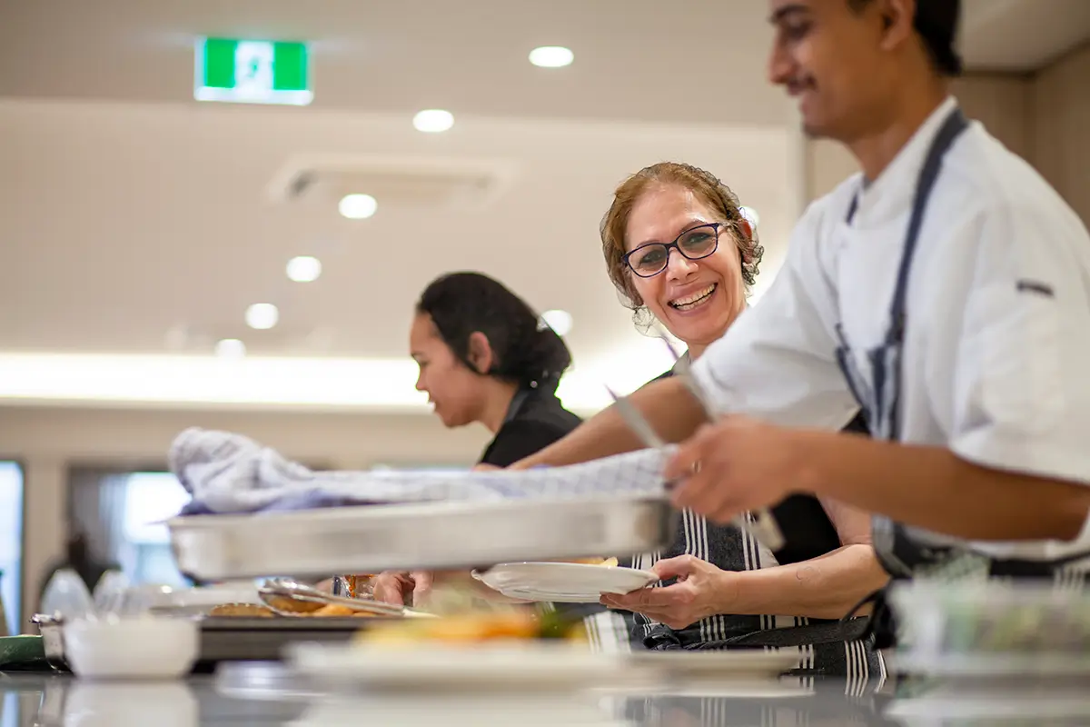 Smiling Italian woman with chefs serving food in a kitchen.