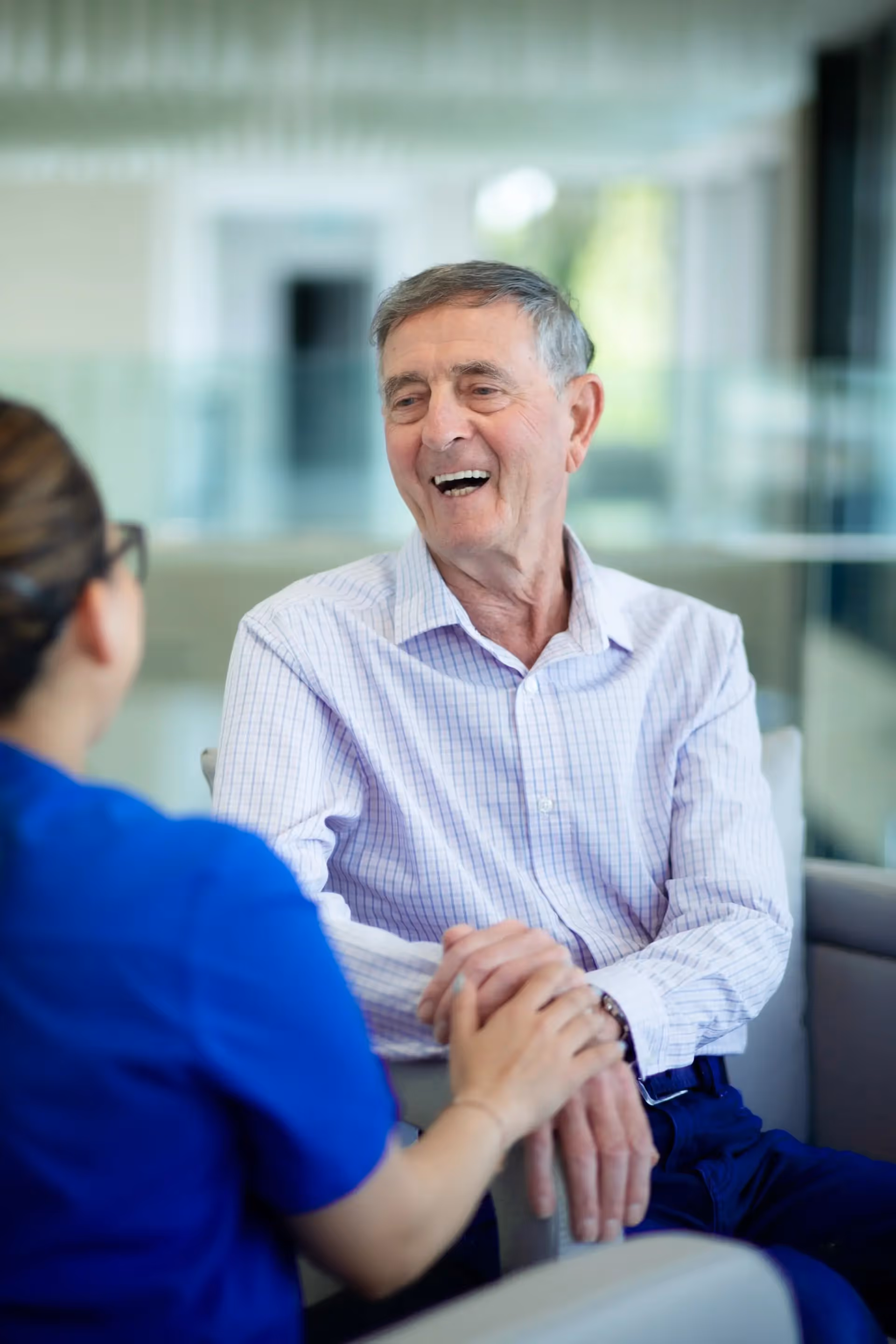 An elderly man in a light checkered shirt smiles while holding hands with a caregiver.