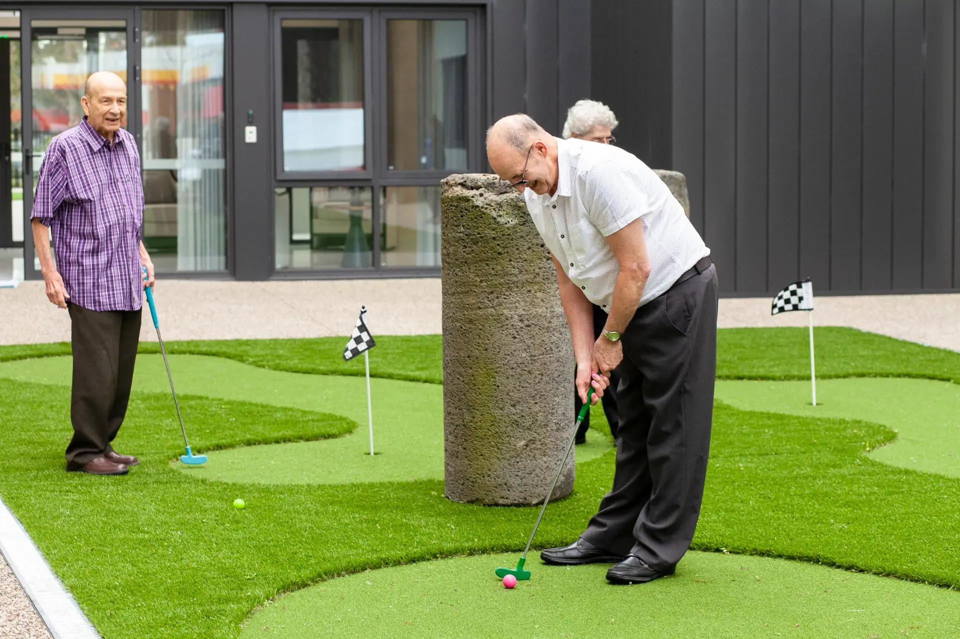 Two elderly men enjoy a game of mini golf on a green outdoor course with small checkered flags, smiling and having fun together