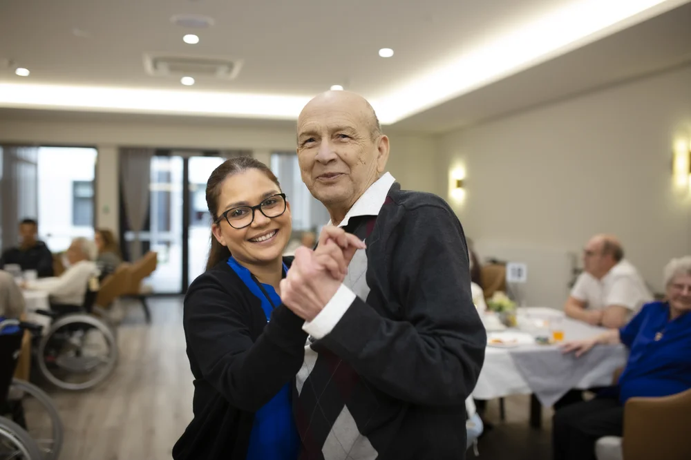 An elderly man and a caregiver smile warmly as they dance together in a bright, welcoming community room.