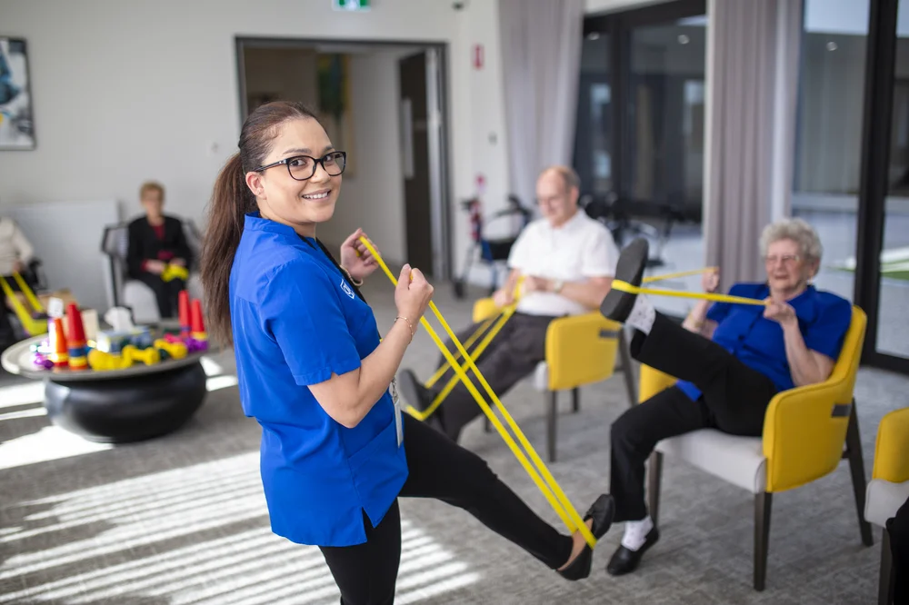 A caregiver in a blue uniform smiles while leading a seated exercise session with resistance bands.