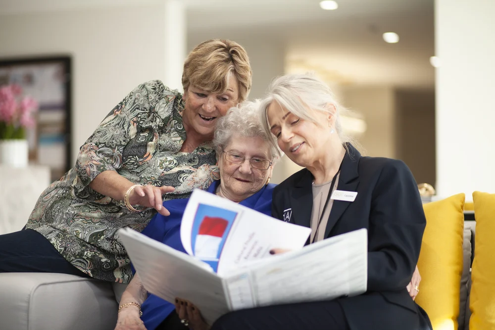 An elderly woman sits with two younger women, all smiling as they look together at documents or a photo album, sharing a warm and engaging moment.