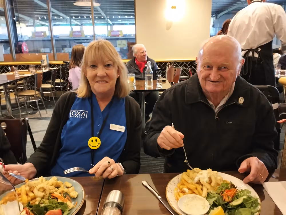 A smiling elderly man and a woman wearing an Oxa Care uniform share a meal at a restaurant, enjoying plates of Greek-style food