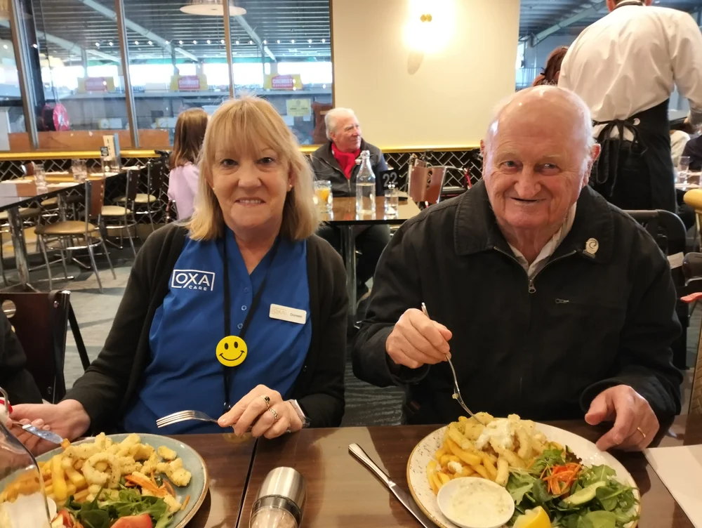 A smiling elderly man and a woman wearing an Oxa Care uniform share a meal at a restaurant, enjoying plates of Greek-style food