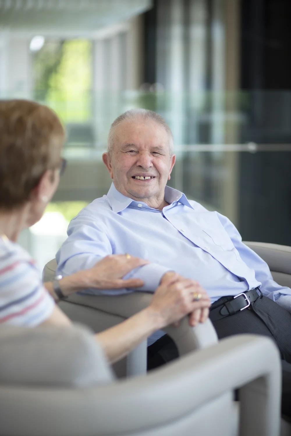 An elderly man in a light blue shirt smiles warmly while sitting in a chair, holding hands with a woman during a friendly conversation.