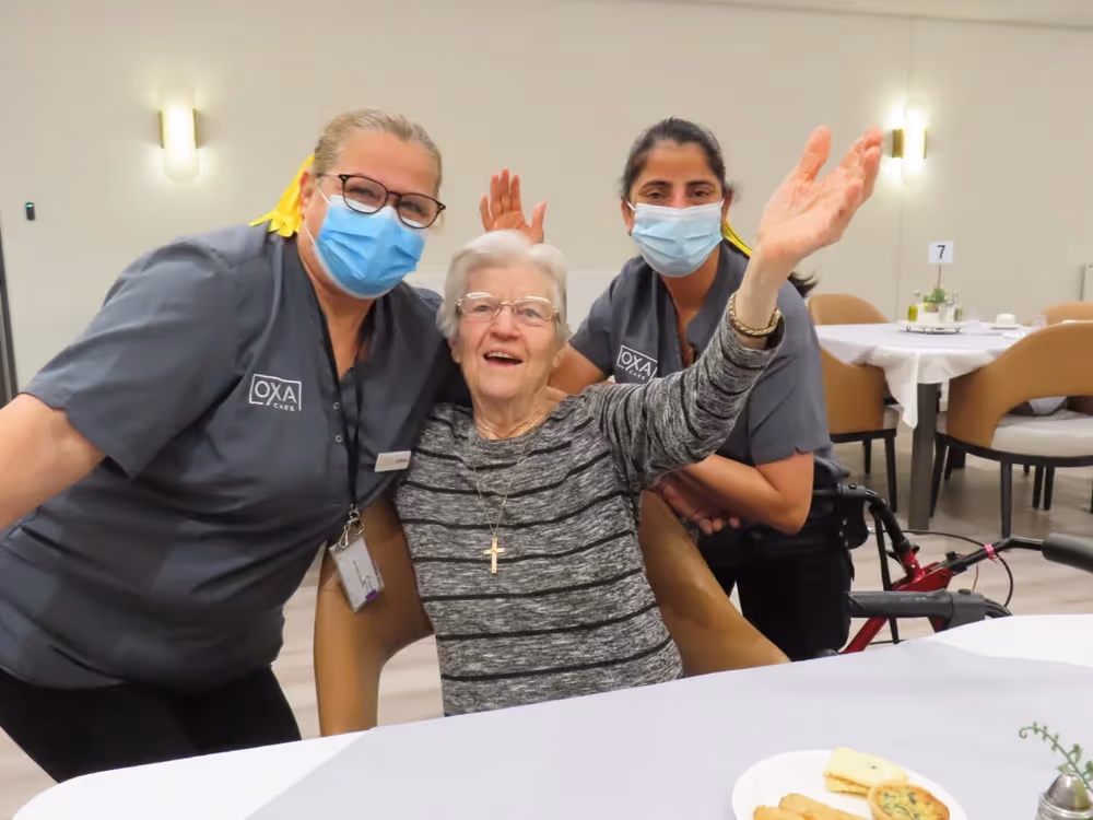 An elderly woman with a big smile raises her arms in joy while sitting between two Oxa Care staff members, all posing happily at a dining table with food.