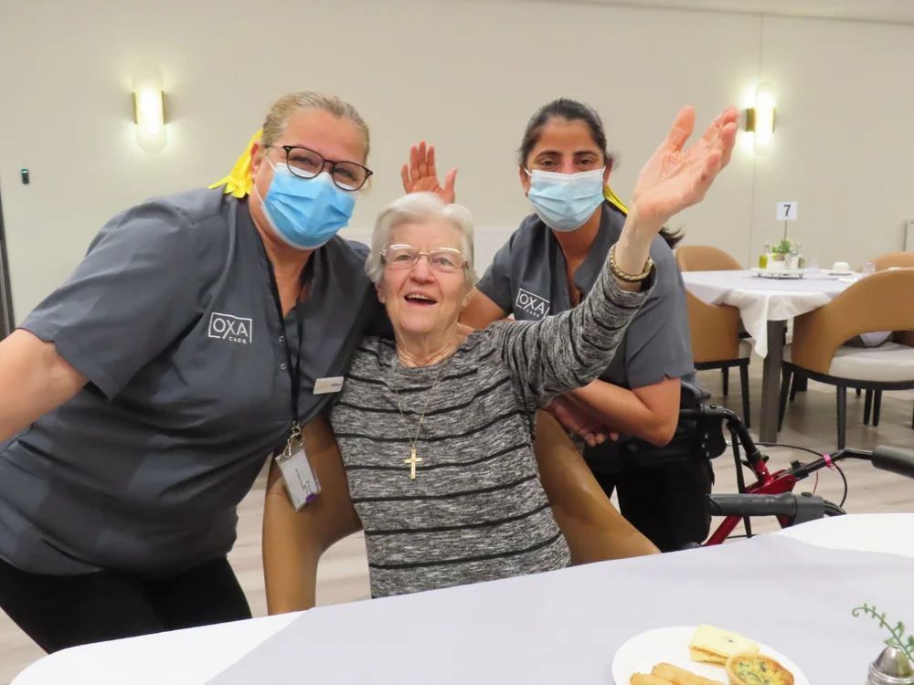 Cheerful elderly Polish woman with arms raised, sitting between two smiling caregivers wearing masks at a dining table.
