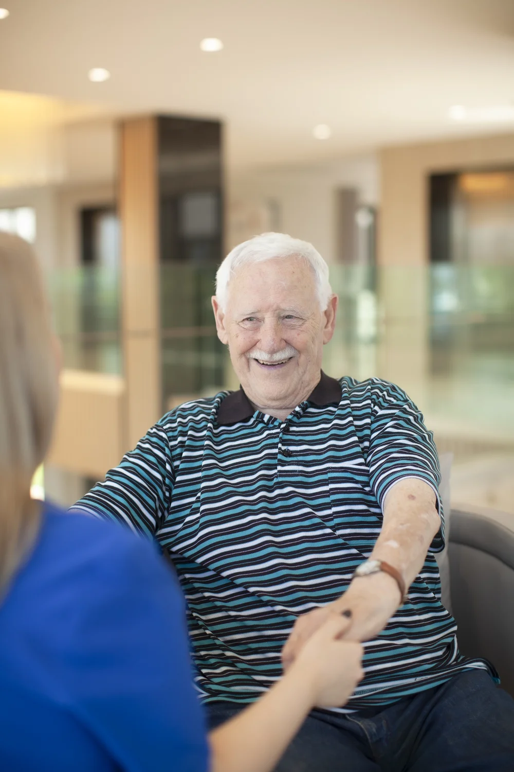 An elderly man with white hair and a striped shirt smiles warmly while holding hands with a woman during a conversation.