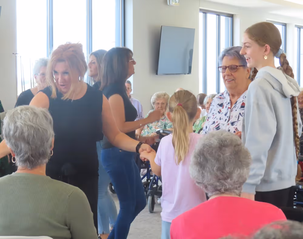 A lively intergenerational gathering where elderly residents, adults, and children smile and hold hands while conversing and dancing indoors.