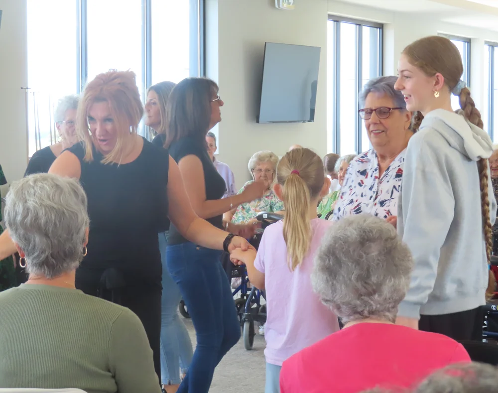 A lively intergenerational gathering where elderly residents, adults, and children smile and hold hands while conversing and dancing indoors.