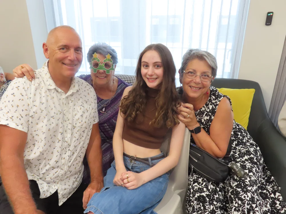 Serbian family group smiling together, including an elderly woman in festive glasses, a young girl, and two adults, during a celebration.