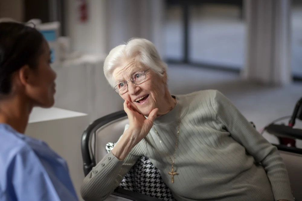 Smiling elderly German woman with white hair and glasses talking to a nurse in a care setting.