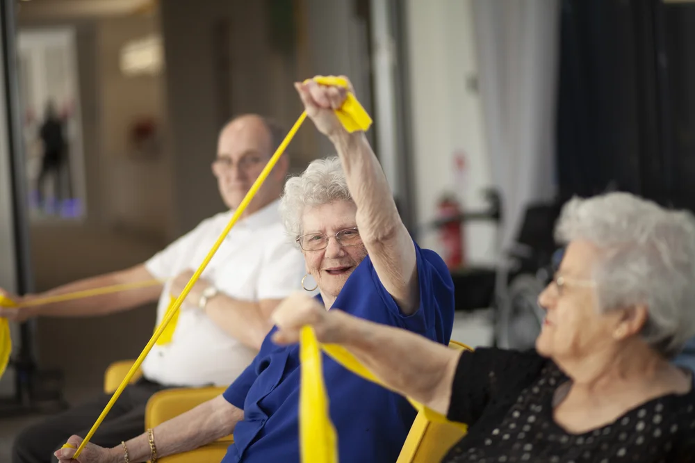 Group of elderly people smiling and exercising with yellow resistance bands in a seated activity session.