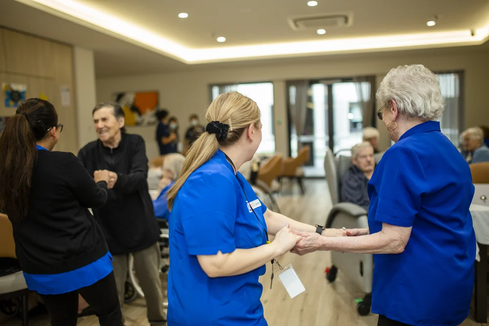 Care staff and elderly residents dancing together in a bright common room.