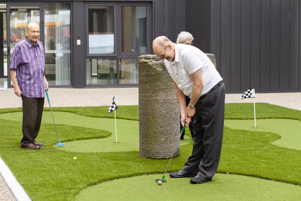 Elderly German men playing mini golf on an outdoor putting green with small checkered flags.