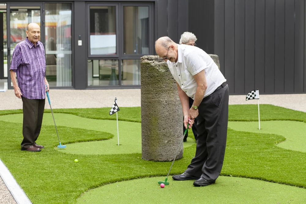 Elderly Serbian men enjoying a game of mini golf on an outdoor putting green with small checkered flags.