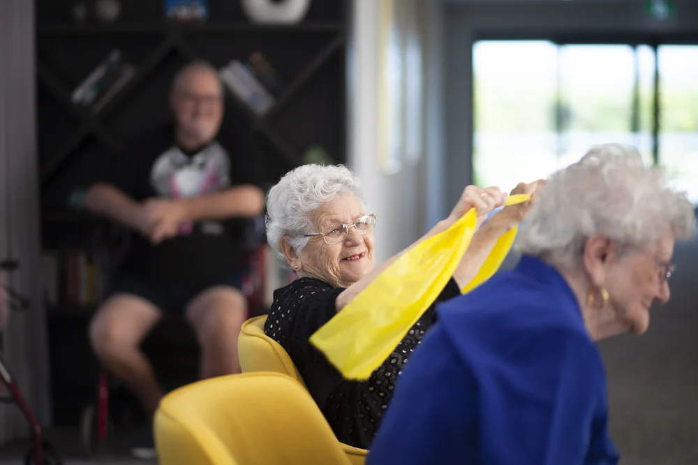 Smiling elderly Polish woman exercising with a yellow resistance band during a group activity session.