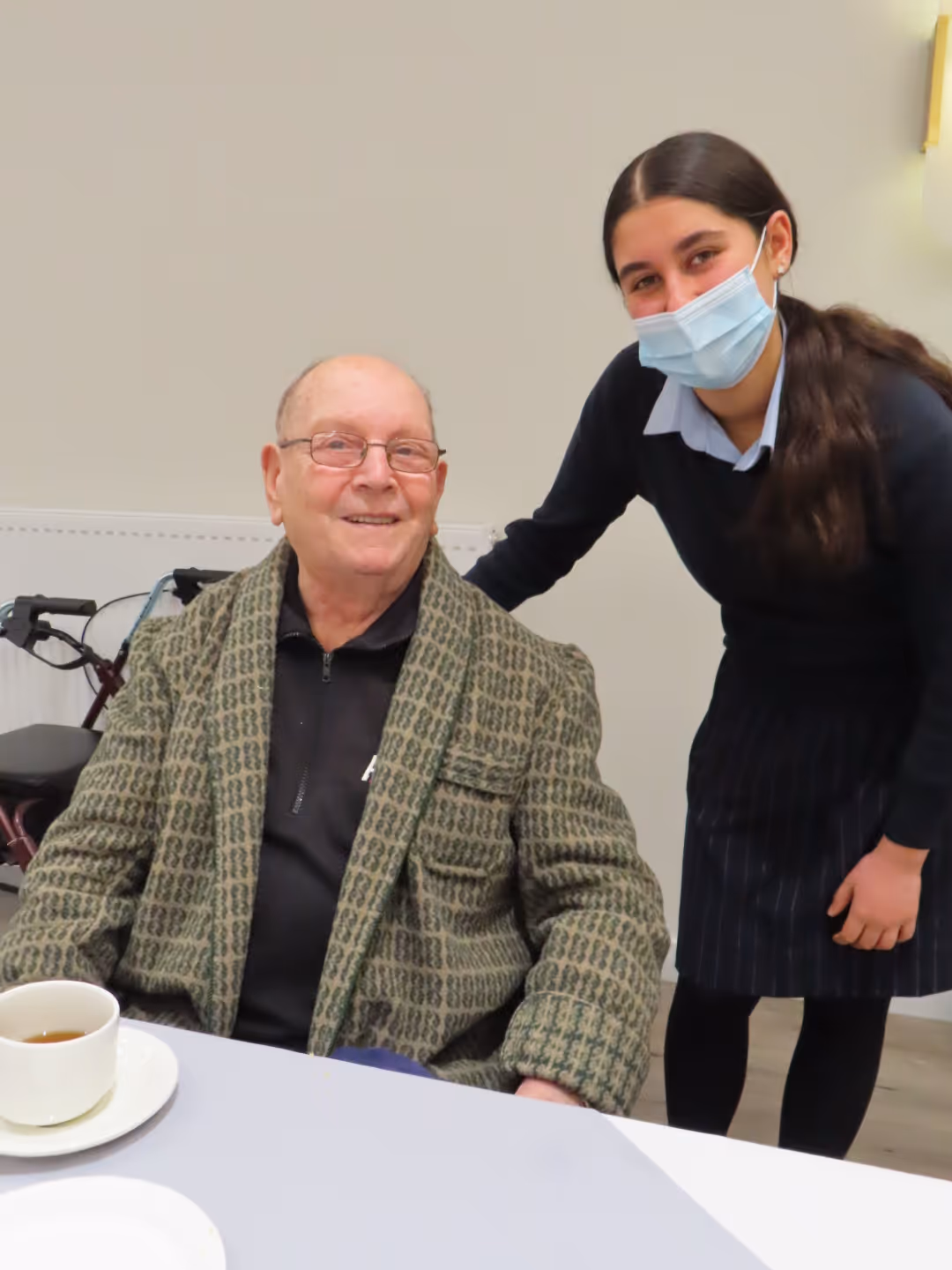 Elderly Polish man sitting at a table with a cup of tea, smiling beside a young woman wearing a face mask.
