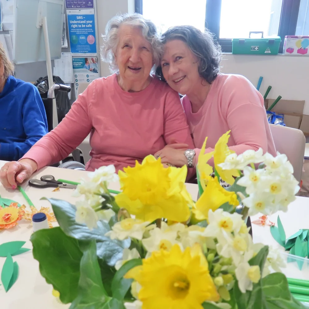 Smiling elderly Polish woman sitting beside another woman in matching pink tops, with bright yellow and white flowers in the foreground.