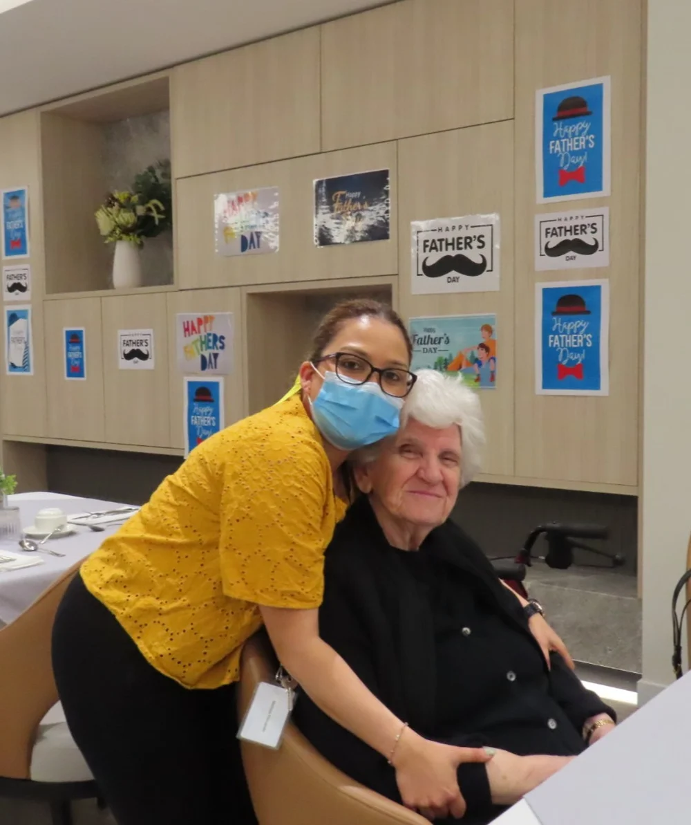 Smiling elderly Russian woman sitting in a chair, embraced by a caregiver.