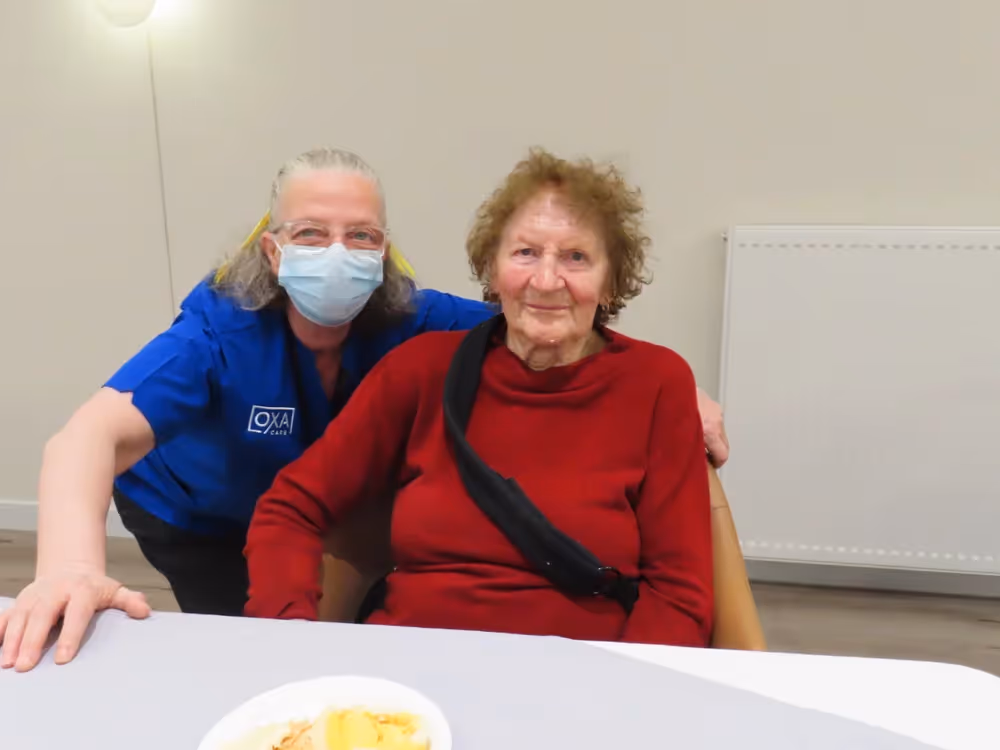 Elderly Russian woman in a red sweater sitting at a table with dessert, smiling beside a caregiver in a blue uniform and mask.